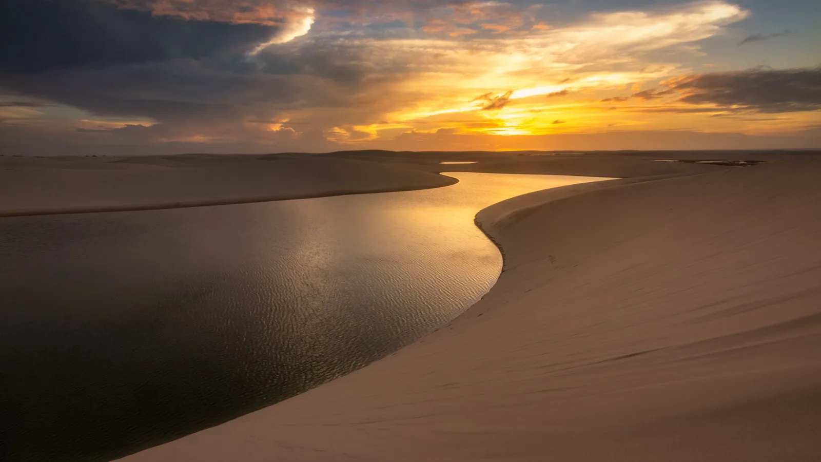 A long lagoon winding away between white dunes at sunset, the water lit gold by a low sun under a sky of dark scattered cloud