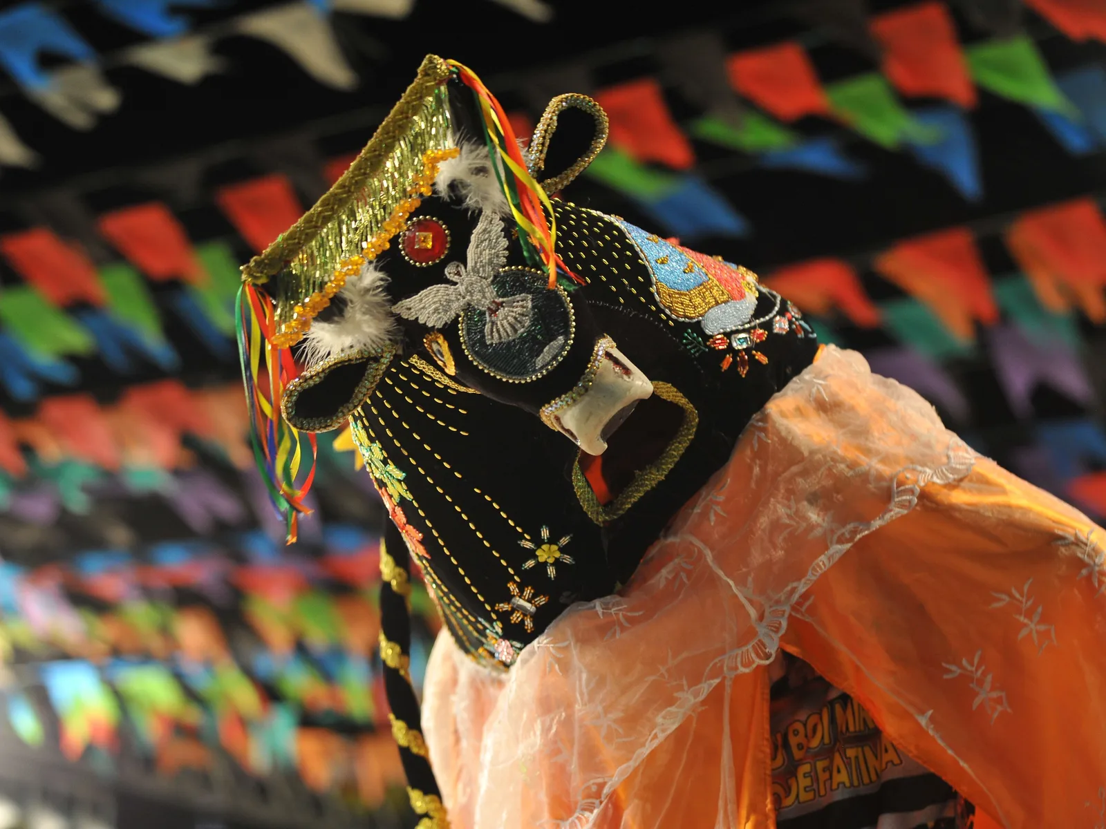The head and shoulders of a bumba meu boi ox costume in close-up, black velvet covered in dense gold, red and green beadwork and sequins, with festival bunting blurred behind it