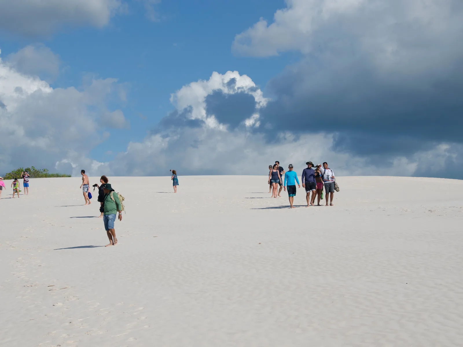 Several separate groups of visitors spread out across a wide white dune under a cloudy sky, walking in loose lines toward the horizon