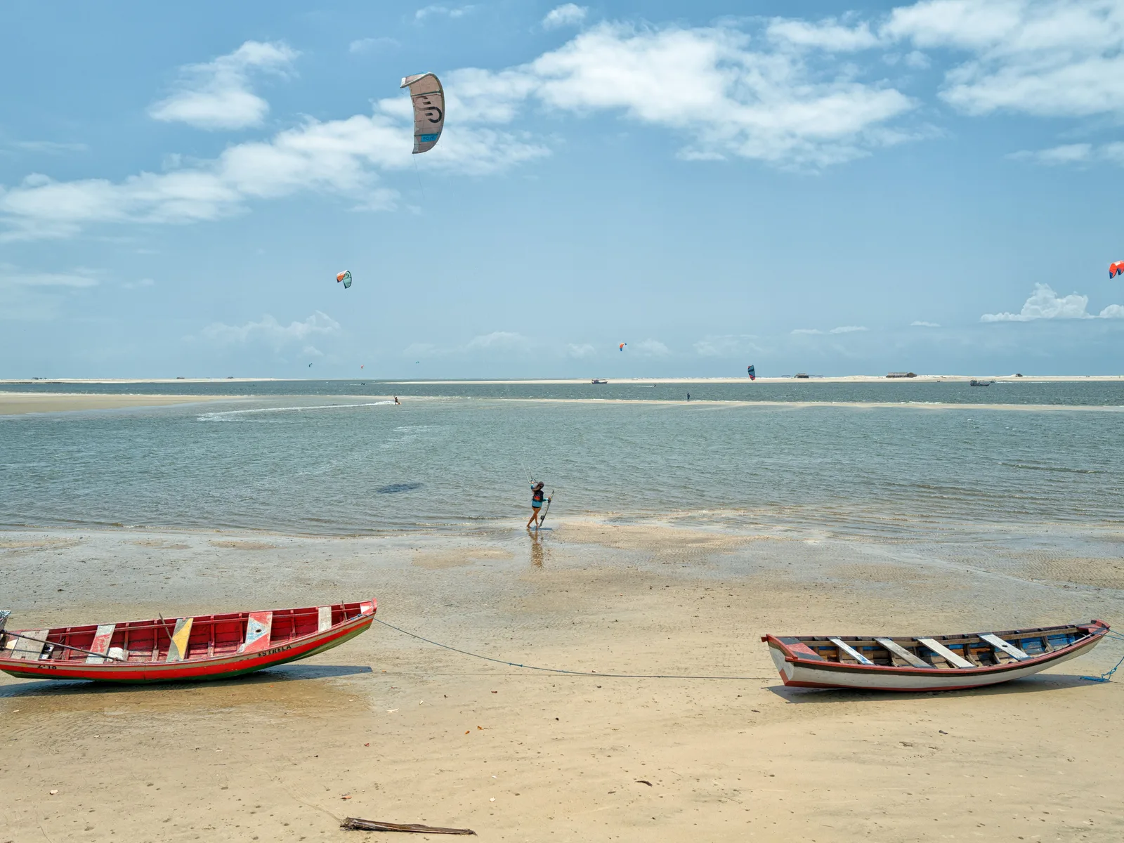 Two wooden fishing boats drawn up on the sand at Atins with kites flying over the flat water behind them and a rider out on the sandbar