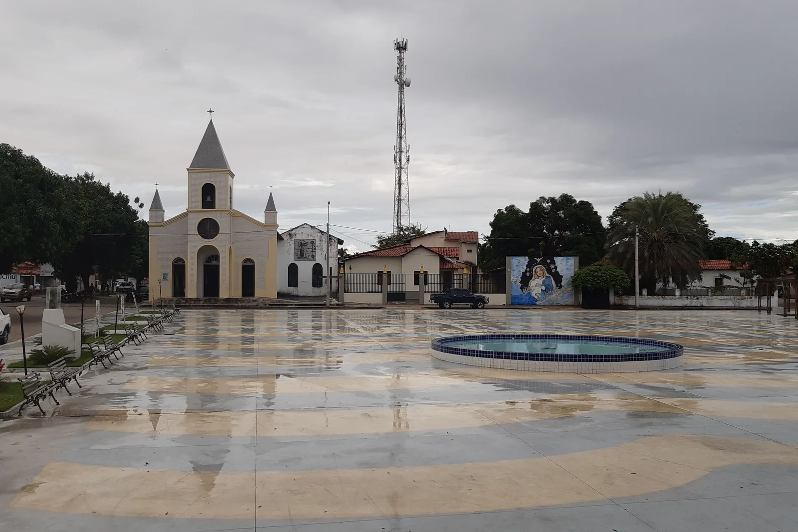 The church and the fountain on the main square in Barreirinhas after rain, the paving still wet and reflecting the gray sky