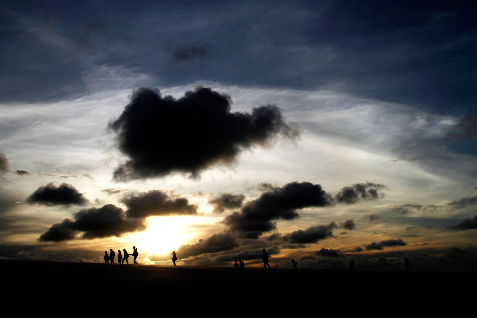 A line of visitors silhouetted on a dune crest at sunset, a heavy dark cloud hanging over the low sun behind them