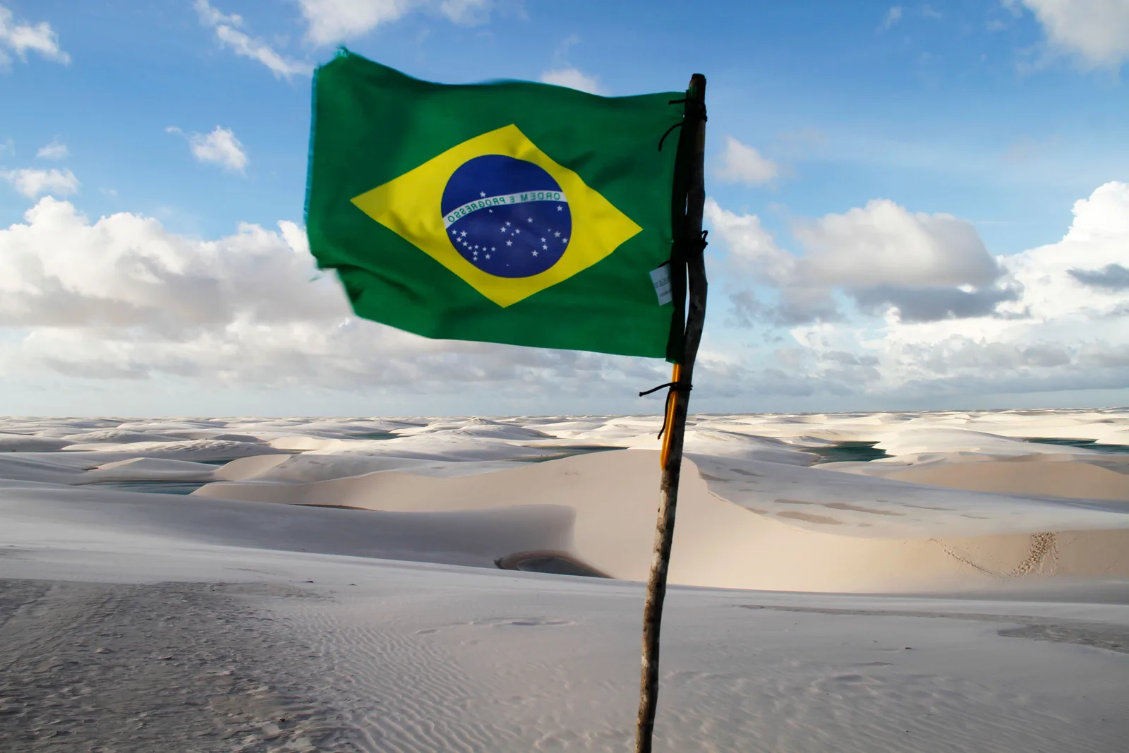 A Brazilian flag flying from a bare wooden pole planted in the sand, with white dunes and a lagoon behind it