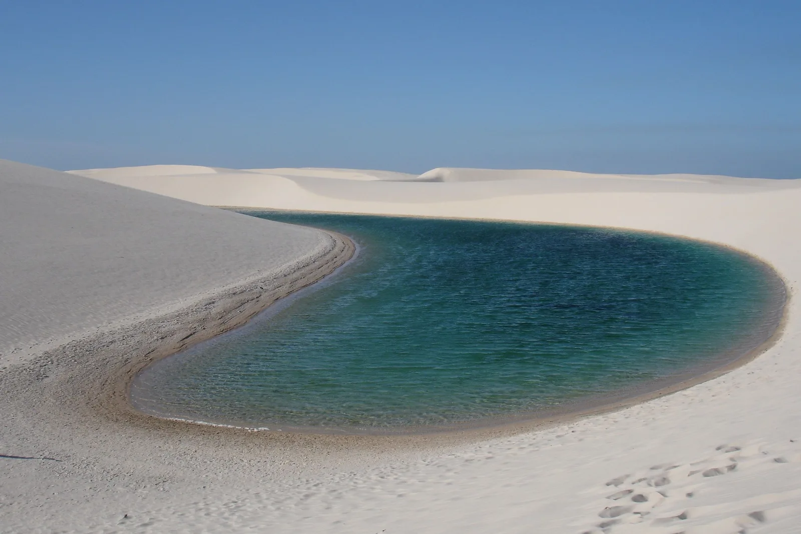 An oval lagoon of clear green water held between two white dunes, footprints crossing the sand toward it