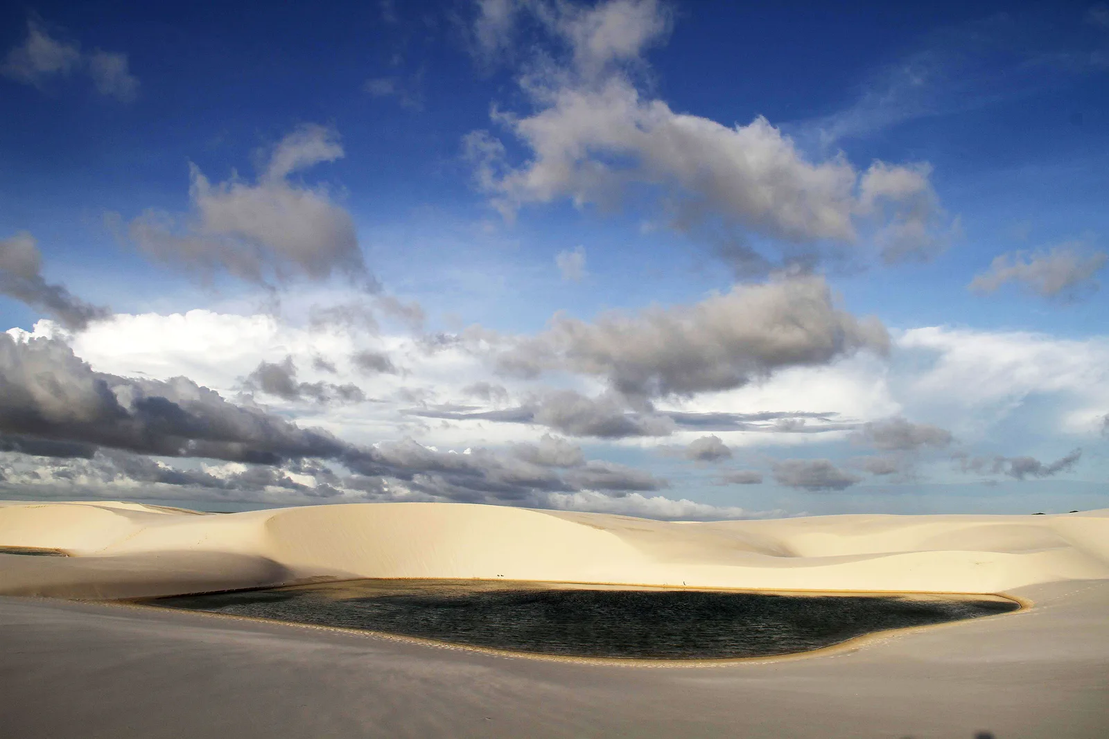 A long bank of gray cloud lying low over the dune field under a deep blue sky, with a broad dark lagoon spread across the sand below it