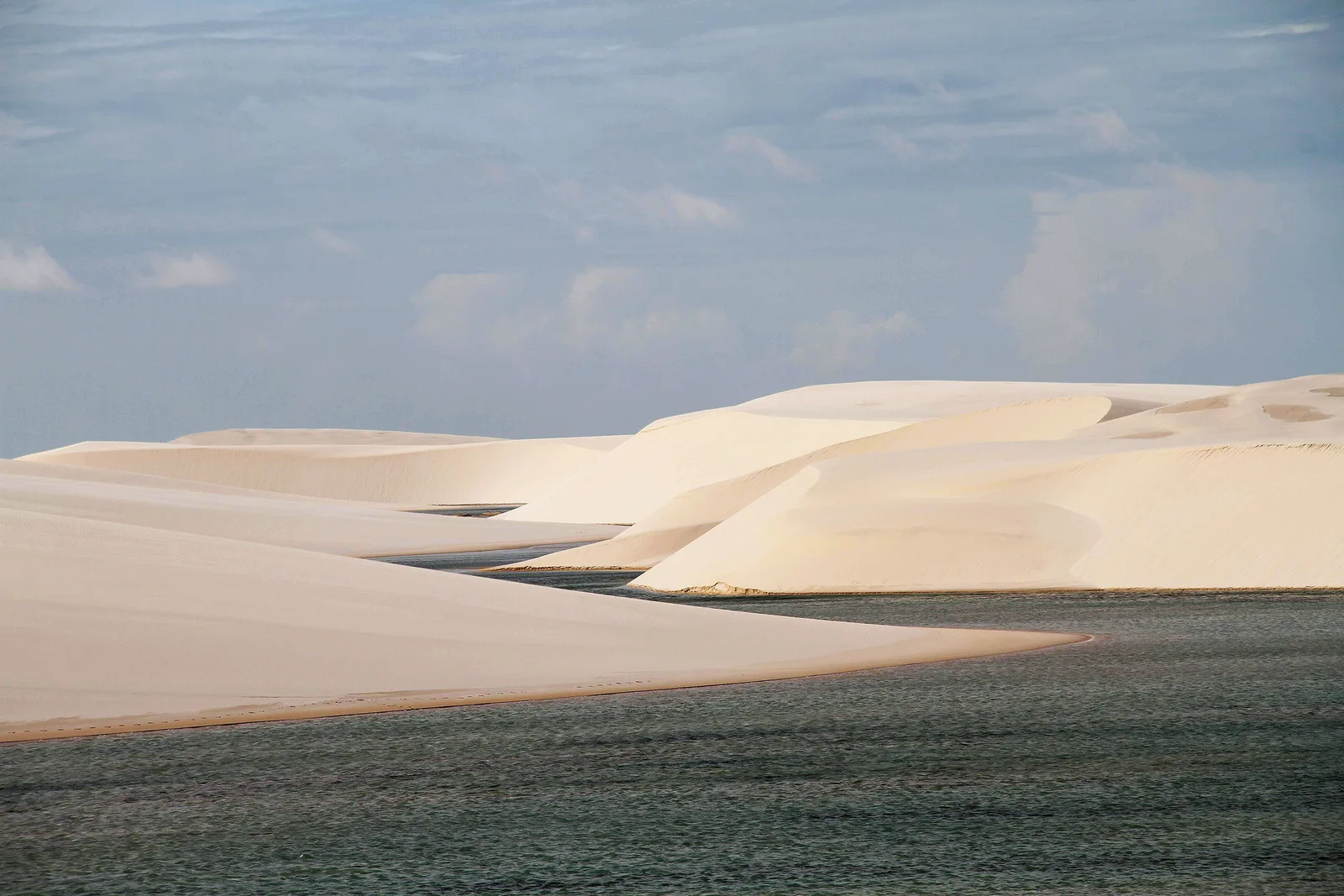 Two long pale lagoons lying either side of a white dune ridge under a hazy sky, the dune field running away flat behind them