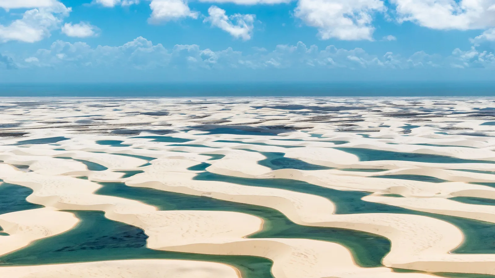 The Lençóis Maranhenses from the air: rank after rank of white sand dunes running to the horizon, with long green and turquoise lagoons lying in every trough between them and the Atlantic as a dark blue band along the top of the frame