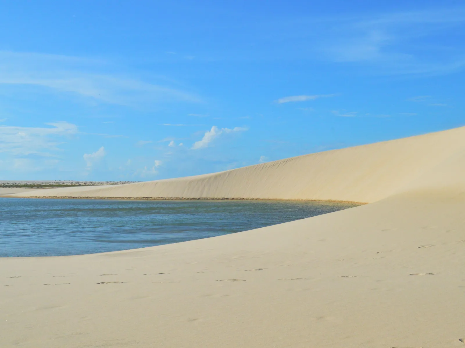 A long smooth white dune slope running down into a broad pale green lagoon near Atins under a bright blue sky, footprints scattered across the sand in the foreground and a low line of dunes on the far horizon