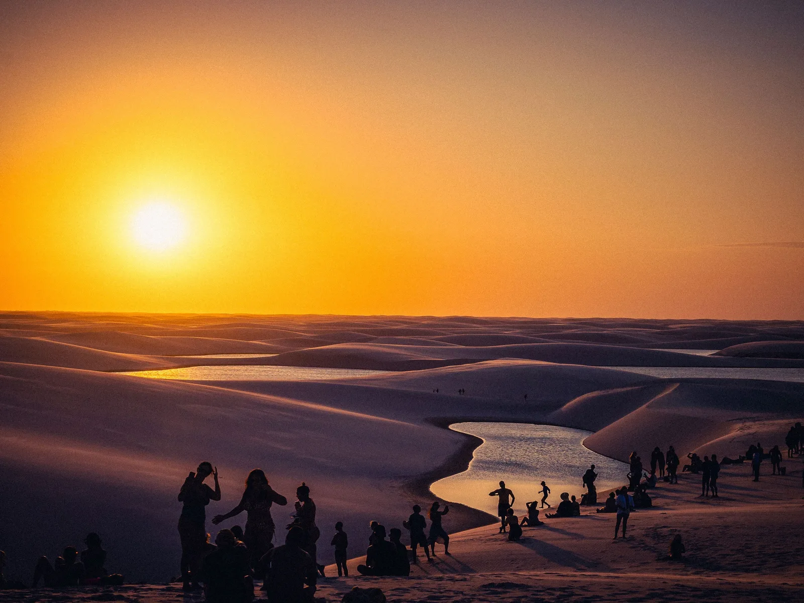 Sunset at Lagoa Bonita: a crowd of visitors in silhouette along a dune above a lagoon, the sun a white disc in an orange sky and birds crossing above them