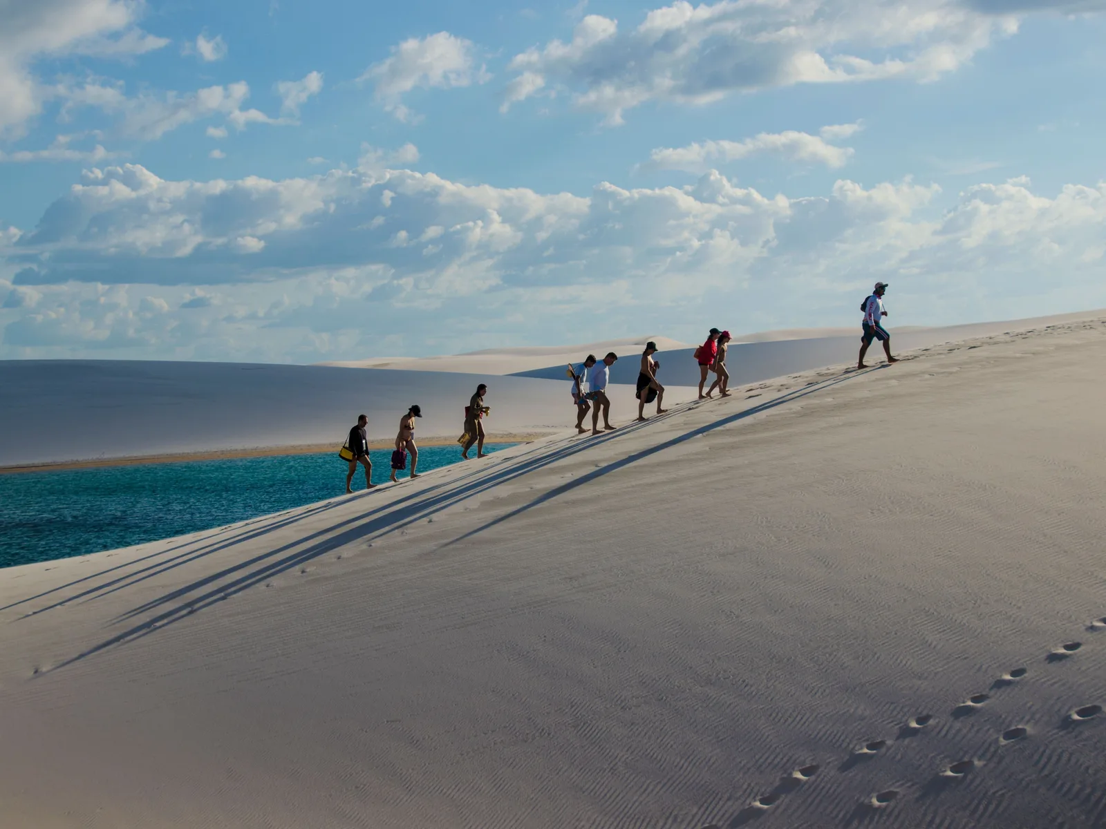 A group walking single file down a dune ridge above a bright turquoise lagoon near the mouth of the Preguiças, vehicle tracks running along the sand beside them