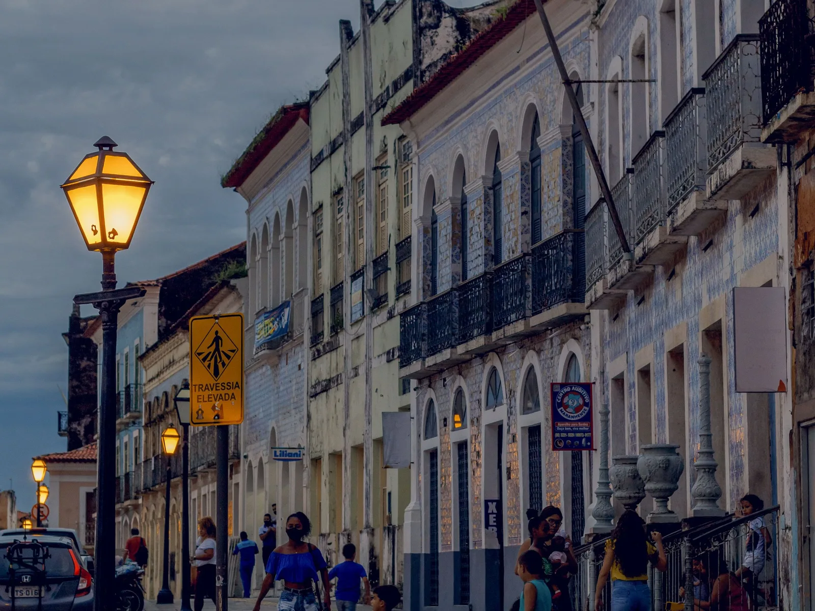 A street in the historic center of São Luís at dusk, a lit iron lamp in the foreground and a row of tall colonial houses running away behind it, their fronts tiled in blue-and-white azulejos above iron balconies, with people walking on the sidewalk below