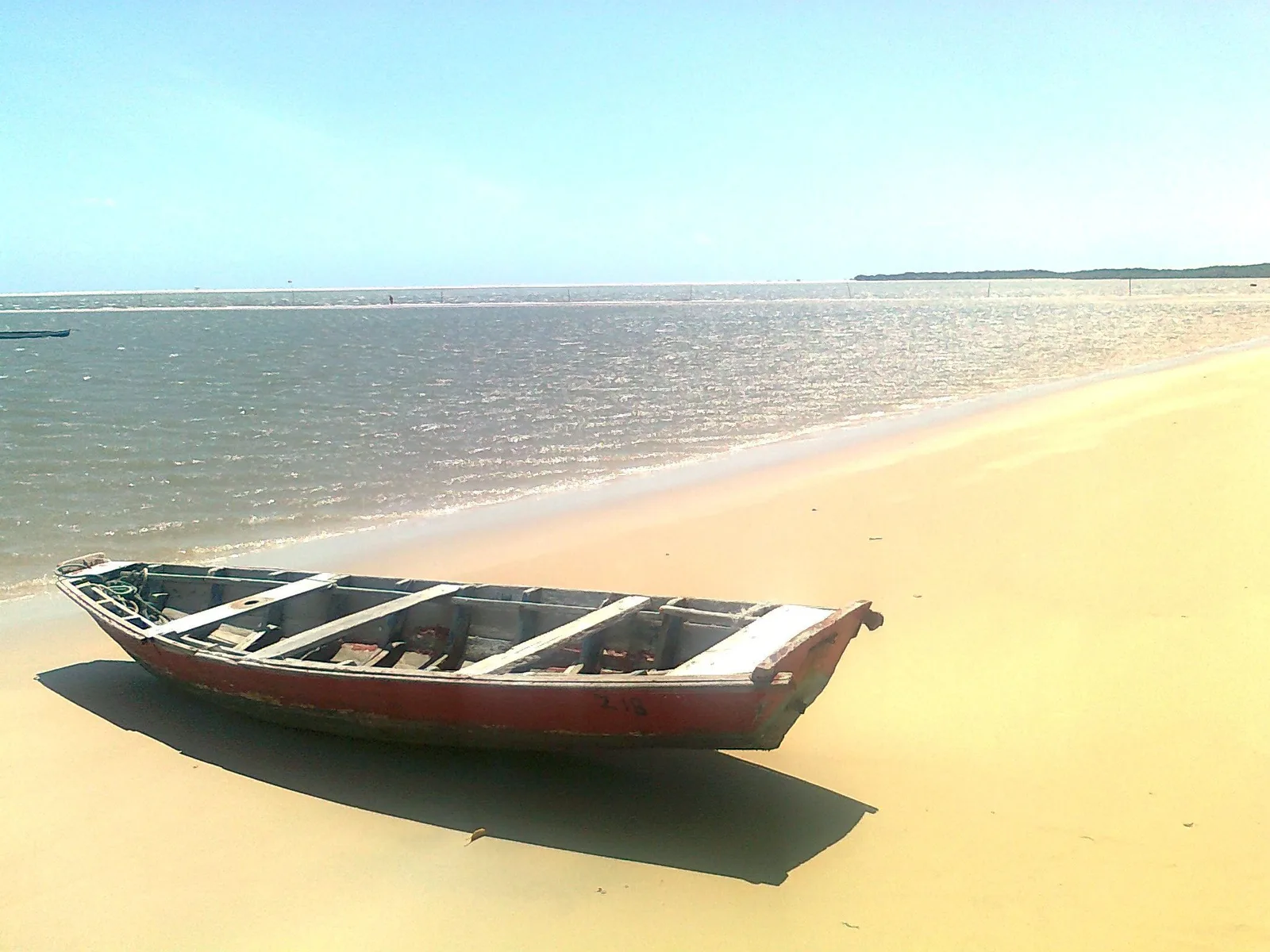 A wooden fishing boat pulled up on the golden sand at the water’s edge at Atins, with flat green water and a low sandbar stretching across the background