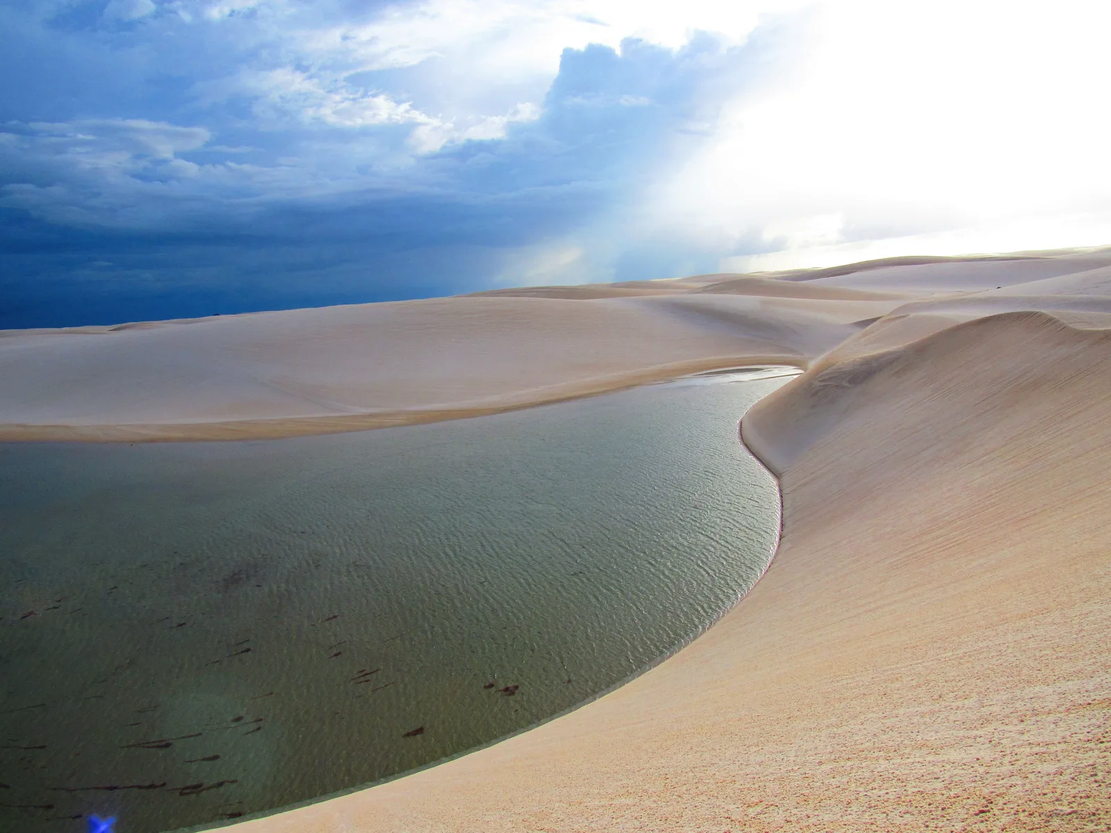 Lagoa Bonita seen from the dune above it in hazy afternoon light: a wide sheet of pale green water curving away between smooth white dunes