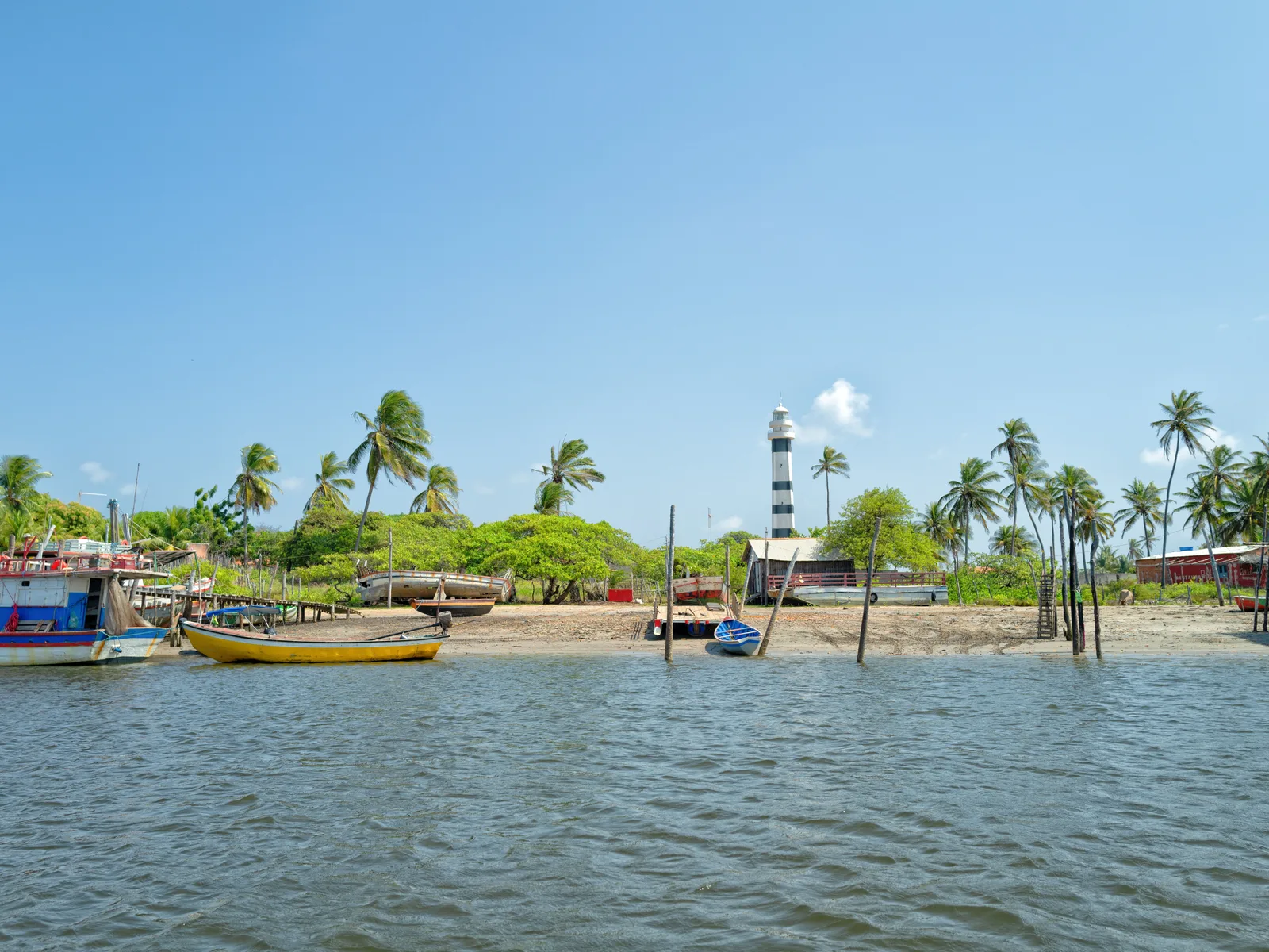 The village of Mandacaru seen from the Rio Preguiças: painted fishing boats moored along a muddy foreshore, palms and low houses behind them, and the black-and-white banded Farol Preguiças rising over the trees