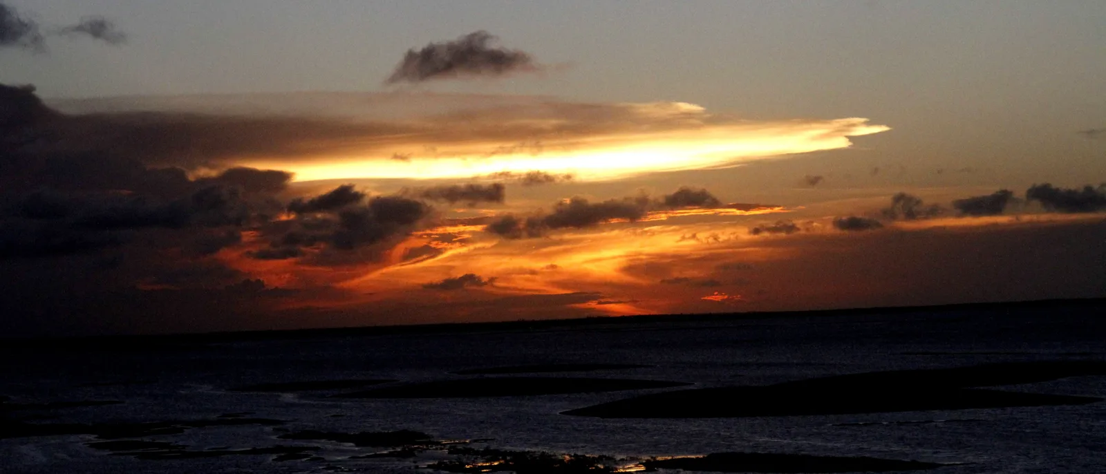 A dramatic orange and gray sunset over dark water near Santo Amaro do Maranhão, a long bar of cloud lit along the horizon
