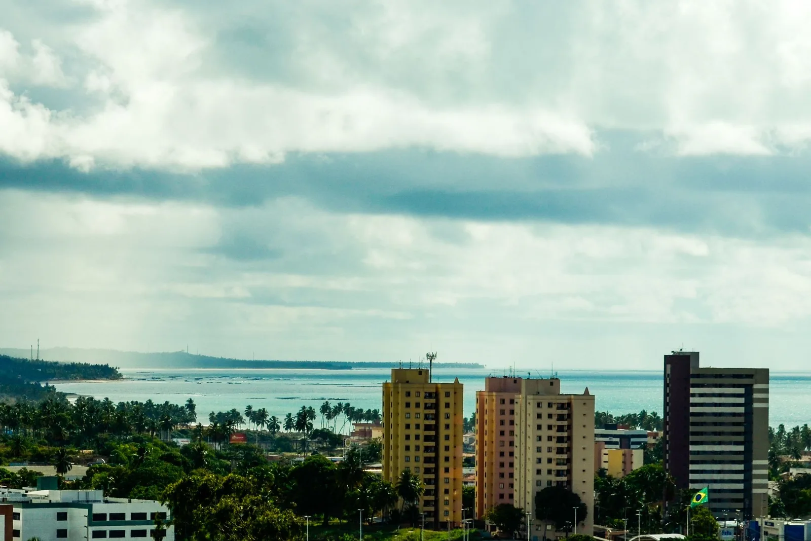 Maceió seen from the higher ground under a heavy sky: apartment blocks and trees in the foreground, and behind them the open ocean with a headland to the left and a long white line of surf breaking on the reef