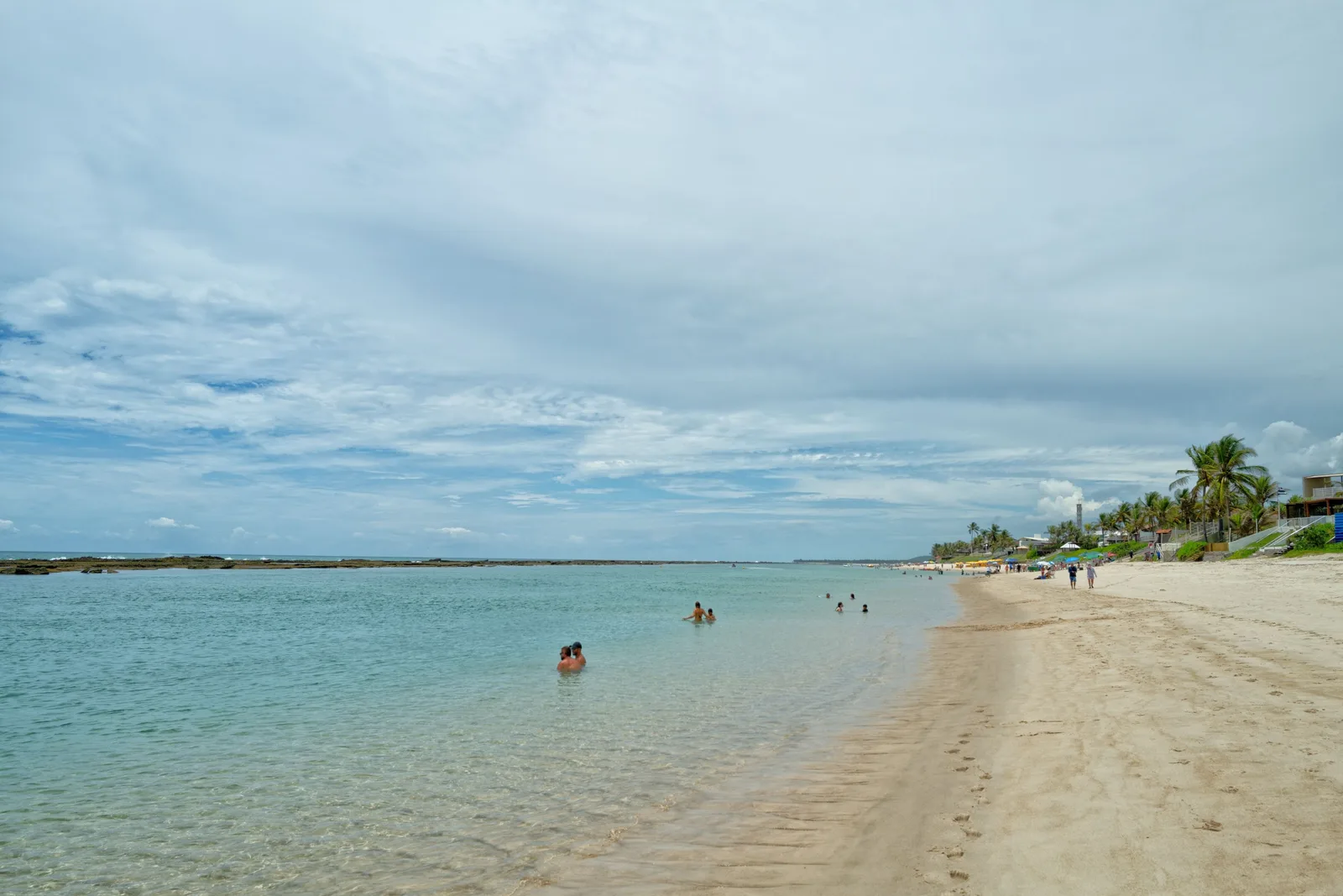 Shallow clear water inside the reef at Praia do Francês with people standing waist-deep in it and a dark reef line breaking the surface further out