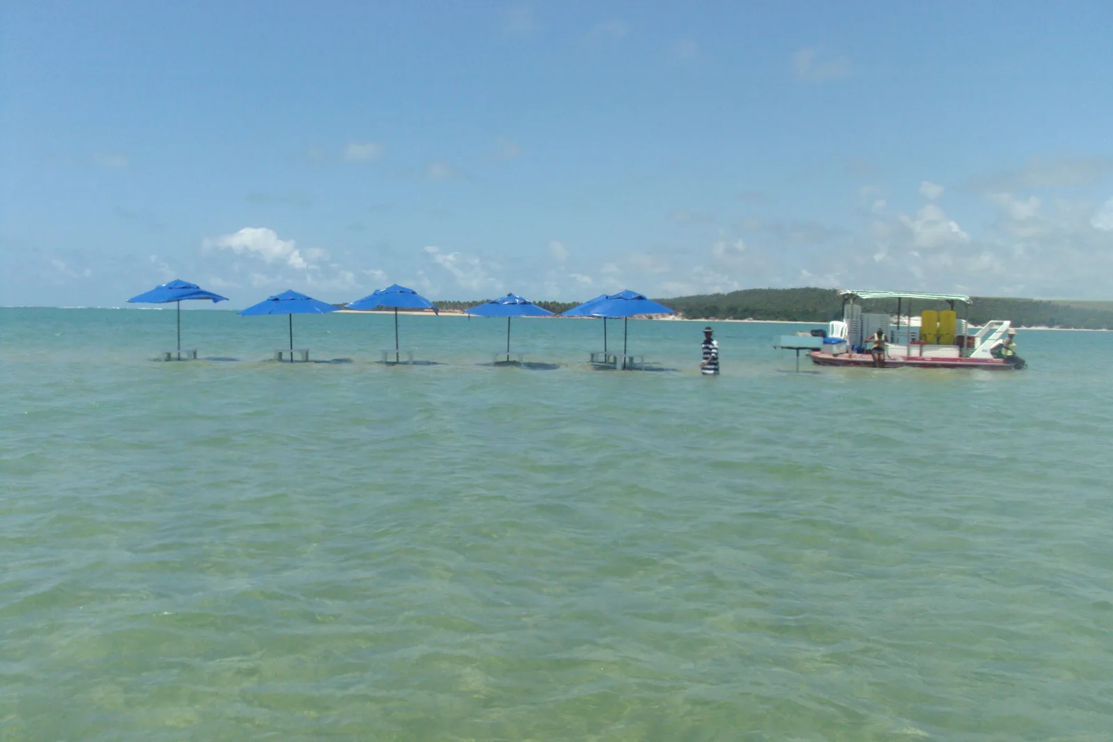Blue parasols standing in shallow water on a sandbank at Praia do Gunga with a small boat moored beyond them and green wooded shore behind