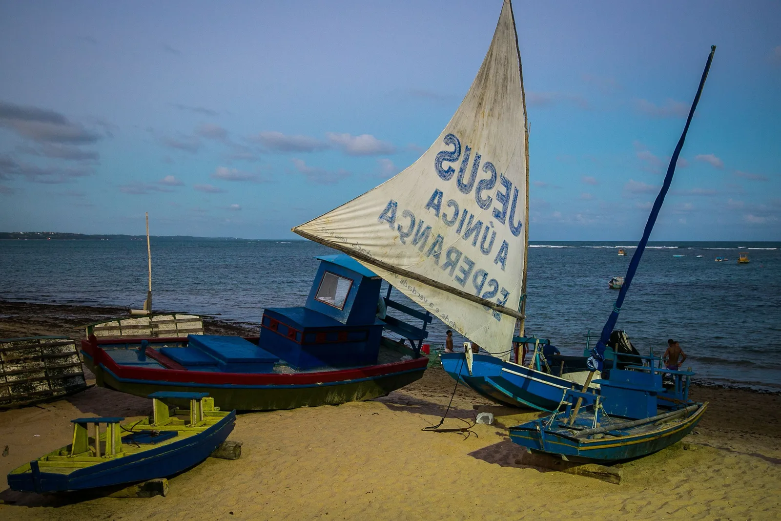A jangada beached at Jatiúca with its sail raised and hand-painted with lettering, three more hulls drawn up around it on the sand and the sea flat behind