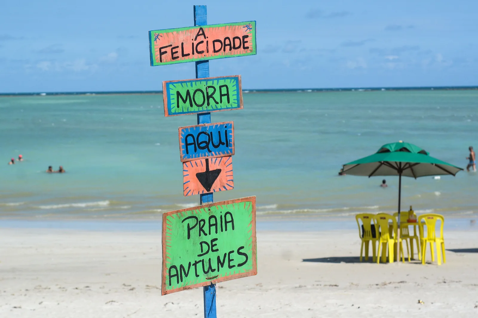 A hand-painted signpost reading Praia de Antunes standing on the sand beside a green parasol and yellow chairs, with calm green water behind