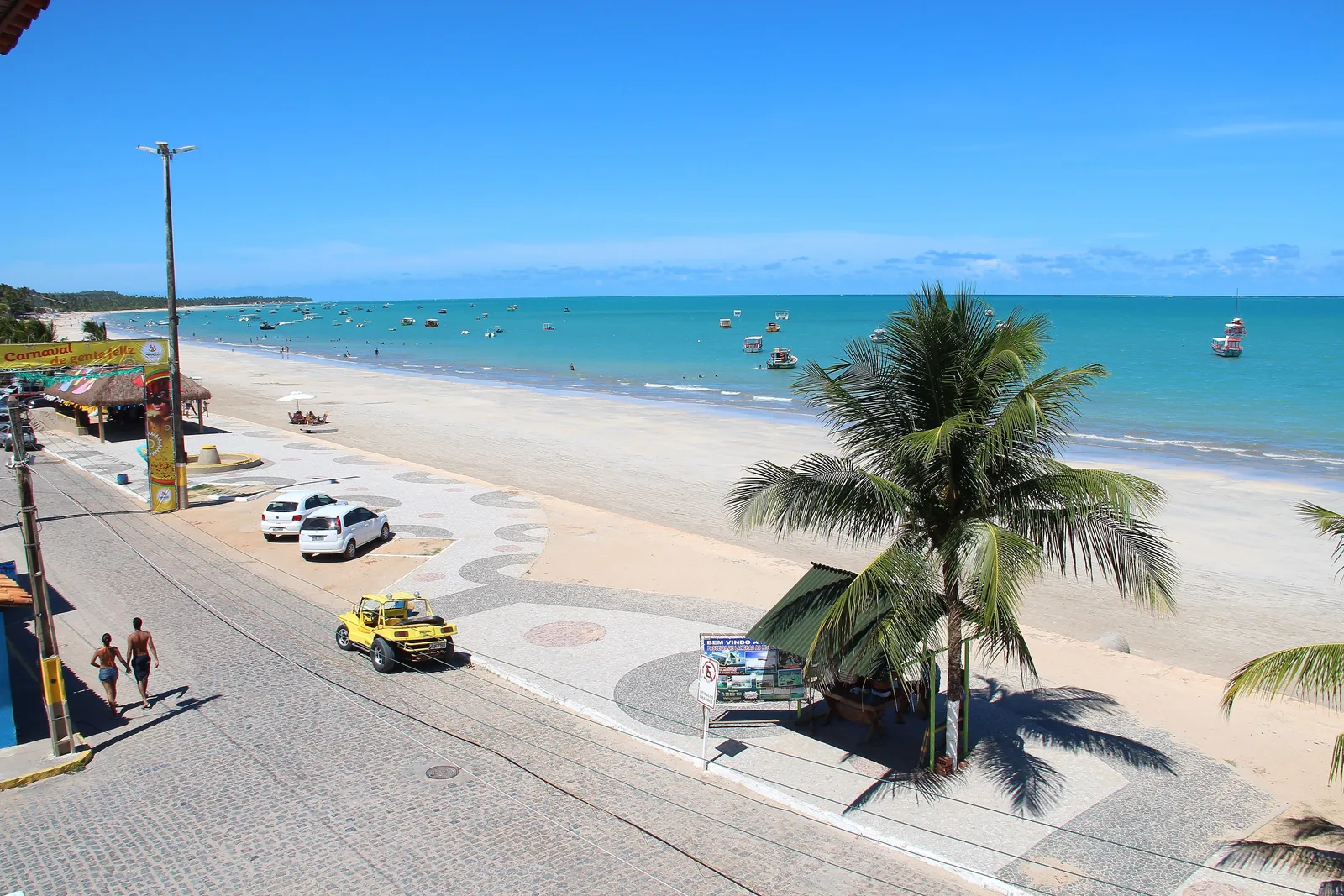 The Maragogi seafront from above: a paved avenue and a patterned promenade with one big coconut palm standing over it, a yellow buggy parked at the curb and fishing boats moored on flat turquoise water offshore