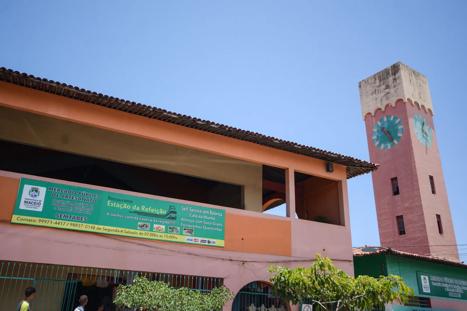 The craft market building in Maceió: a low pink and cream hall with a tall tiled clock tower beside it against a bright blue sky