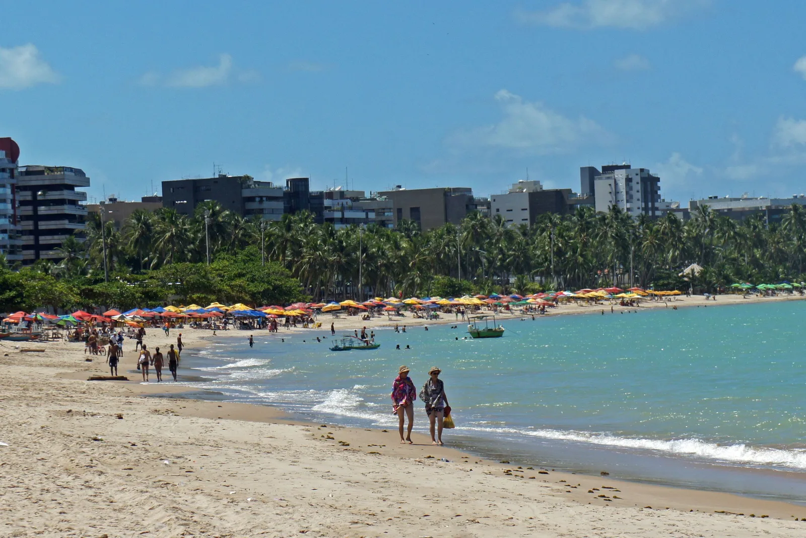 Pajuçara beach in the afternoon: rows of parasols along the sand, people walking at the waterline and the towers of the seafront rising behind the palms