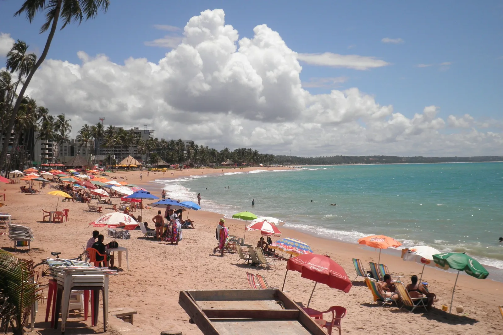 Ponta Verde beach on a busy afternoon: colored parasols and chairs along the sand, people in the shallows and the curve of the bay running away behind