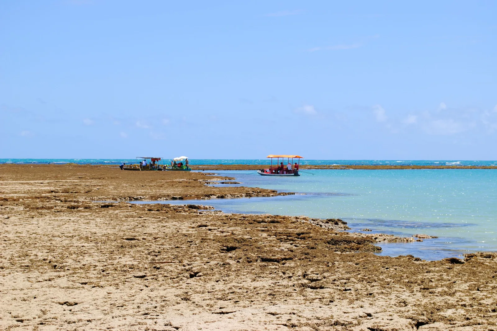 The exposed reef flat at low tide off Praia do Patacho with dark coral and shallow pools in the foreground and two small tour boats anchored out on the water beyond