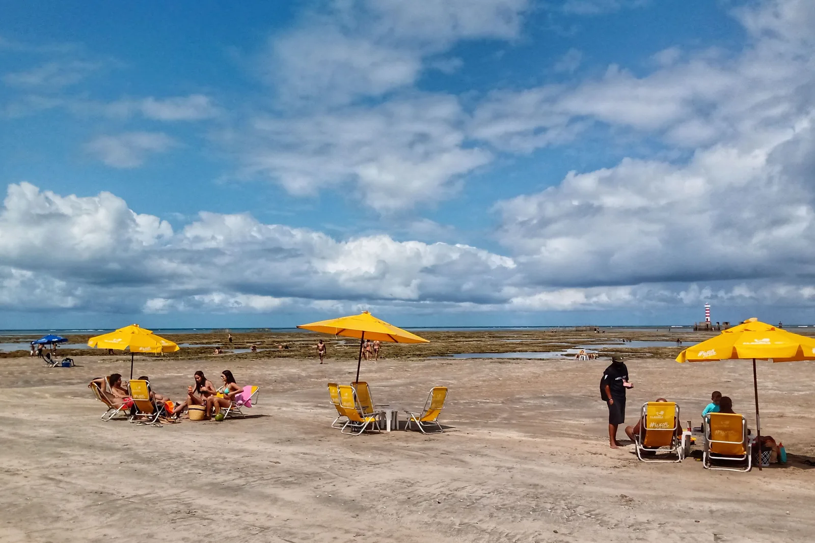 Yellow parasols and beach chairs set out on the wet flats at low tide, people sitting under them on damp sand with the dark exposed reef and the sea further out under a big cloudy sky