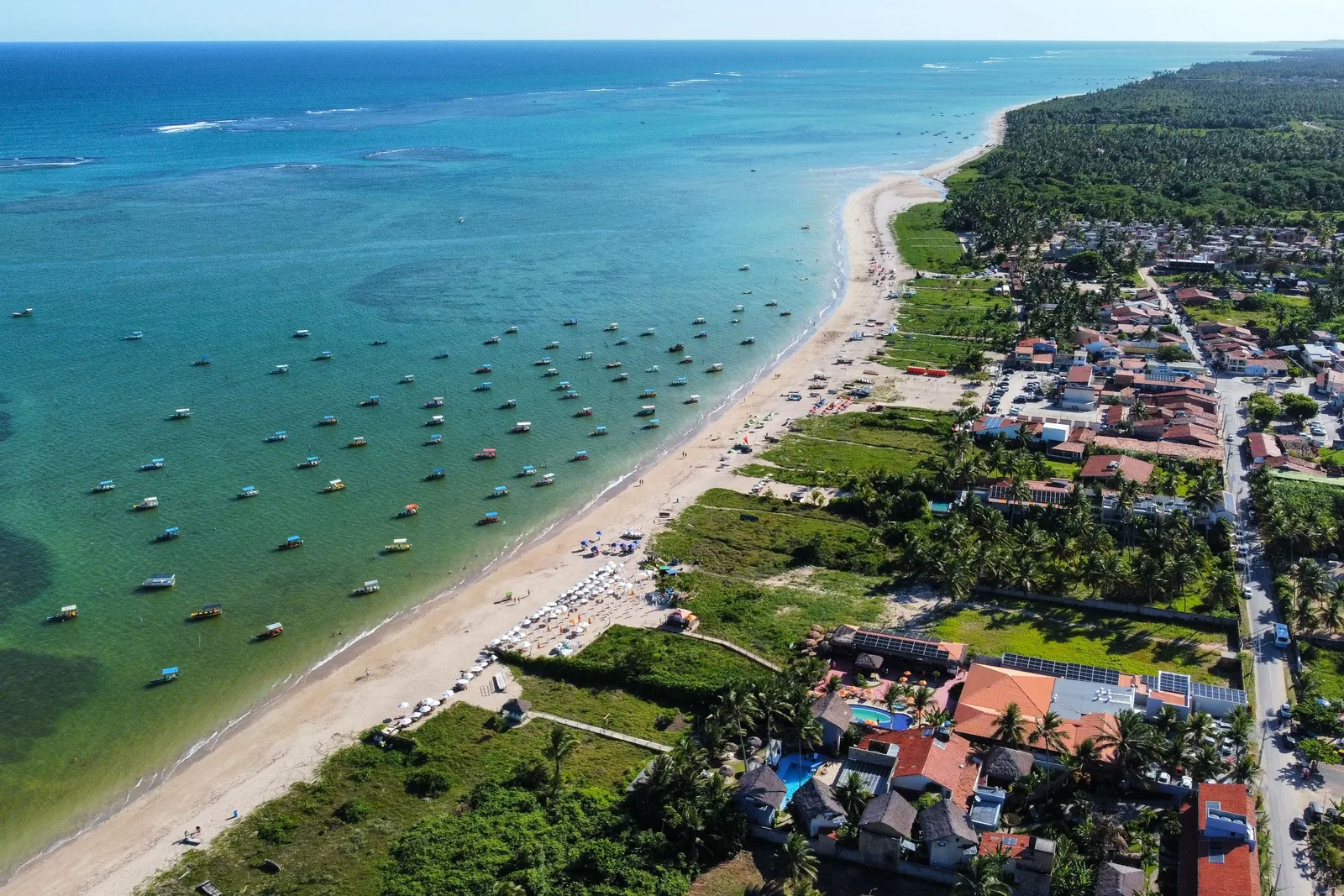 The village of Porto da Rua at São Miguel dos Milagres from the air: red-roofed houses among palms along a pale beach, with small boats moored on turquoise water inside a reef