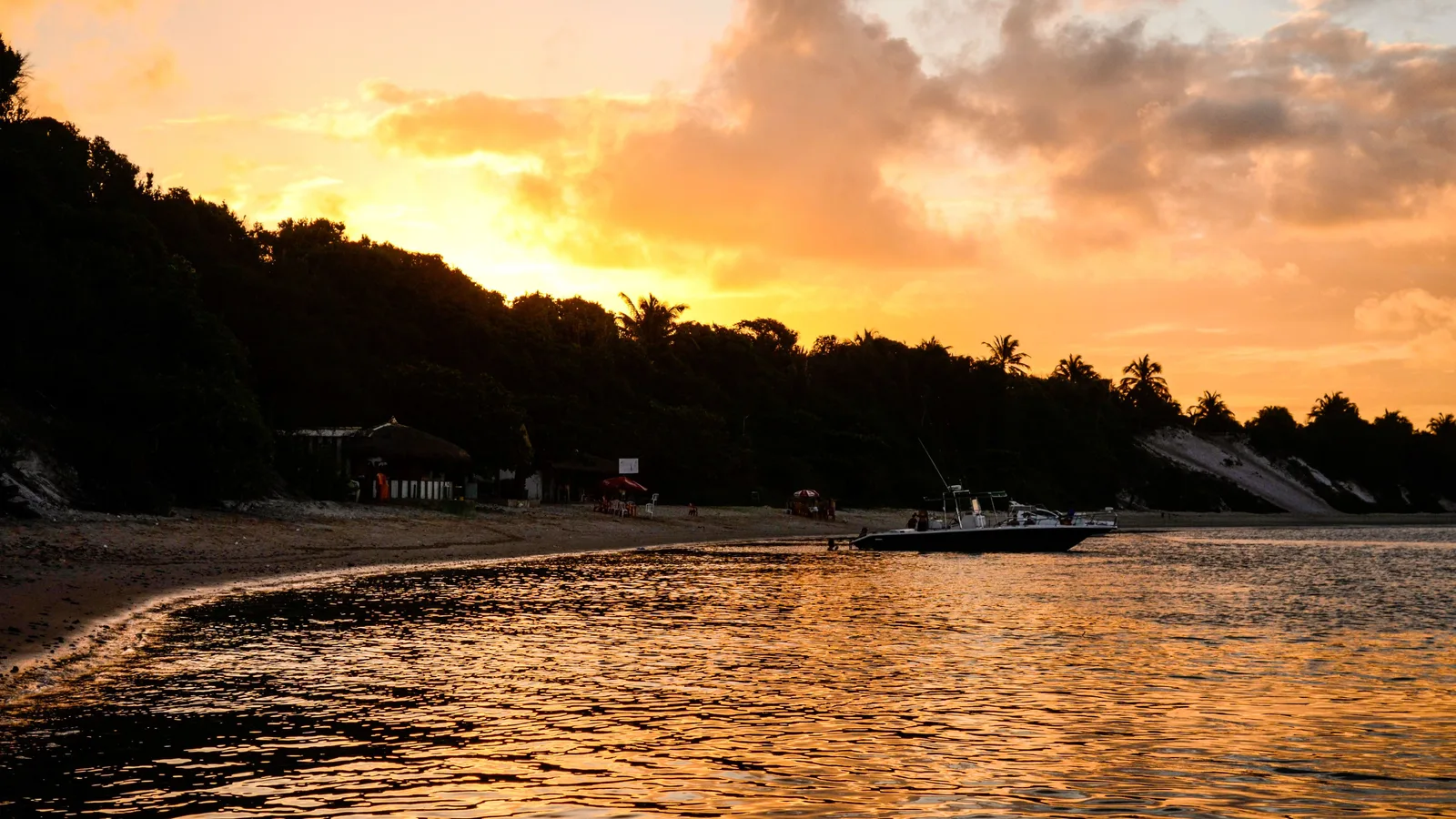 Sunset at Praia do Gunga: the sun low behind a dark line of palms, warm light across the water and a small boat moored offshore