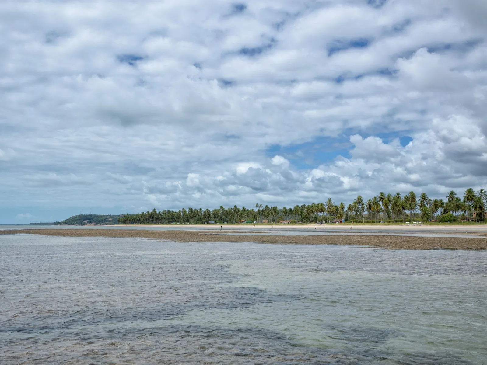 A wide flat beach at low water under a sky packed with white cumulus, shallow water running over pale sand flats and a low line of coconut palms in the distance