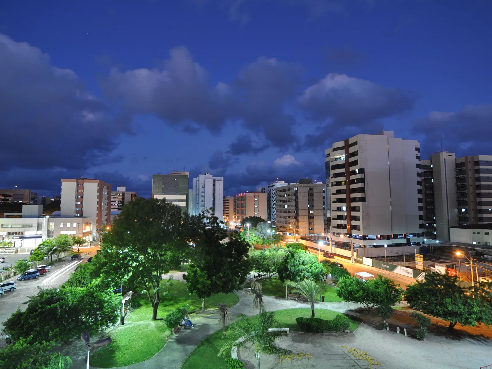 Maceió at dusk from above: apartment blocks along a lit avenue with a floodlit green park of lawns and trees in the foreground under a deep blue sky