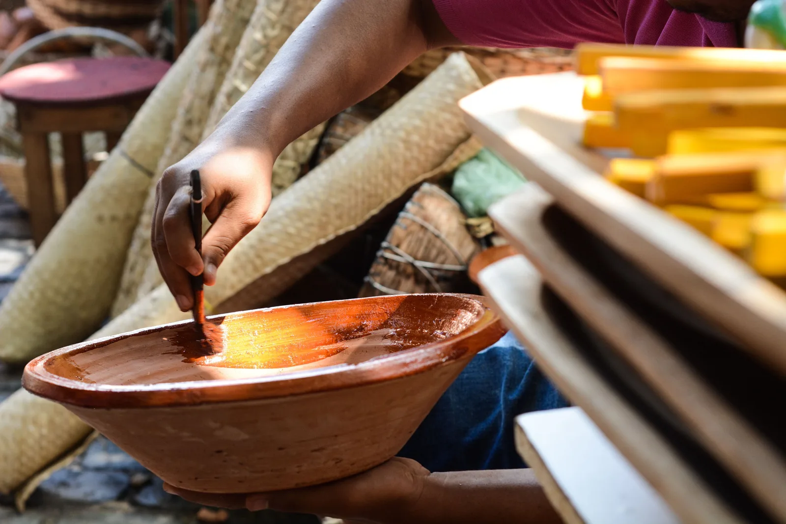 A craftworker brushing varnish into a shallow red clay bowl held on the lap, with rolled straw matting and stacked yellow-painted wood behind