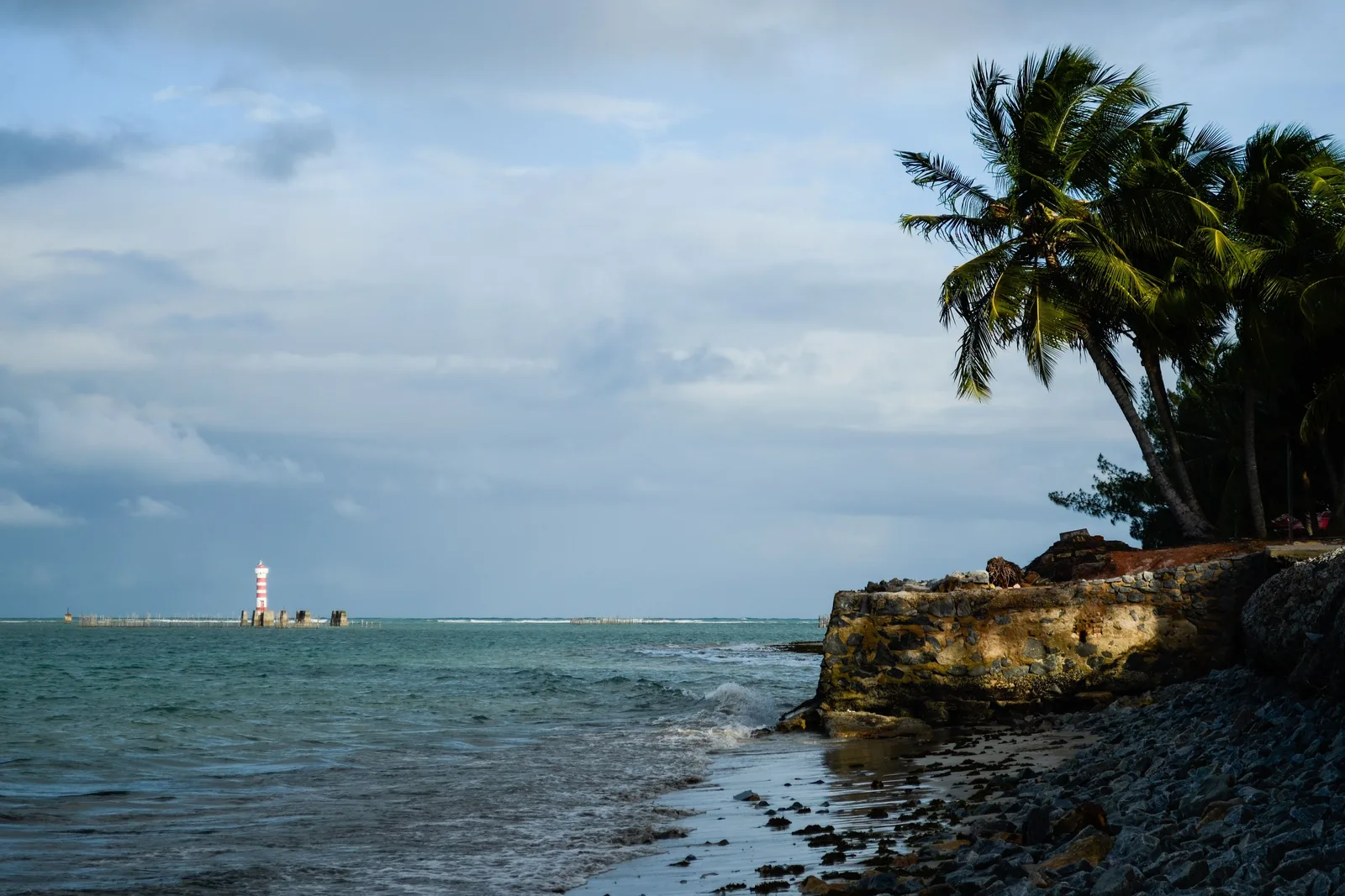 The Ponta Verde lighthouse seen low across the water at low tide, dark rocks and palms in the foreground under a flat gray sky