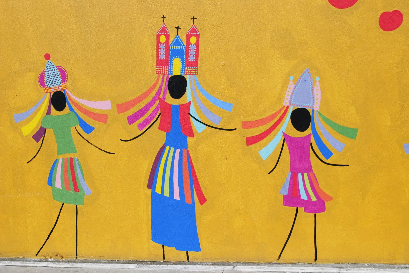 A mural on a yellow wall showing three Alagoan guerreiro figures in striped fringed costumes and colored ribbons, the middle one wearing a pink twin-towered church as a headdress