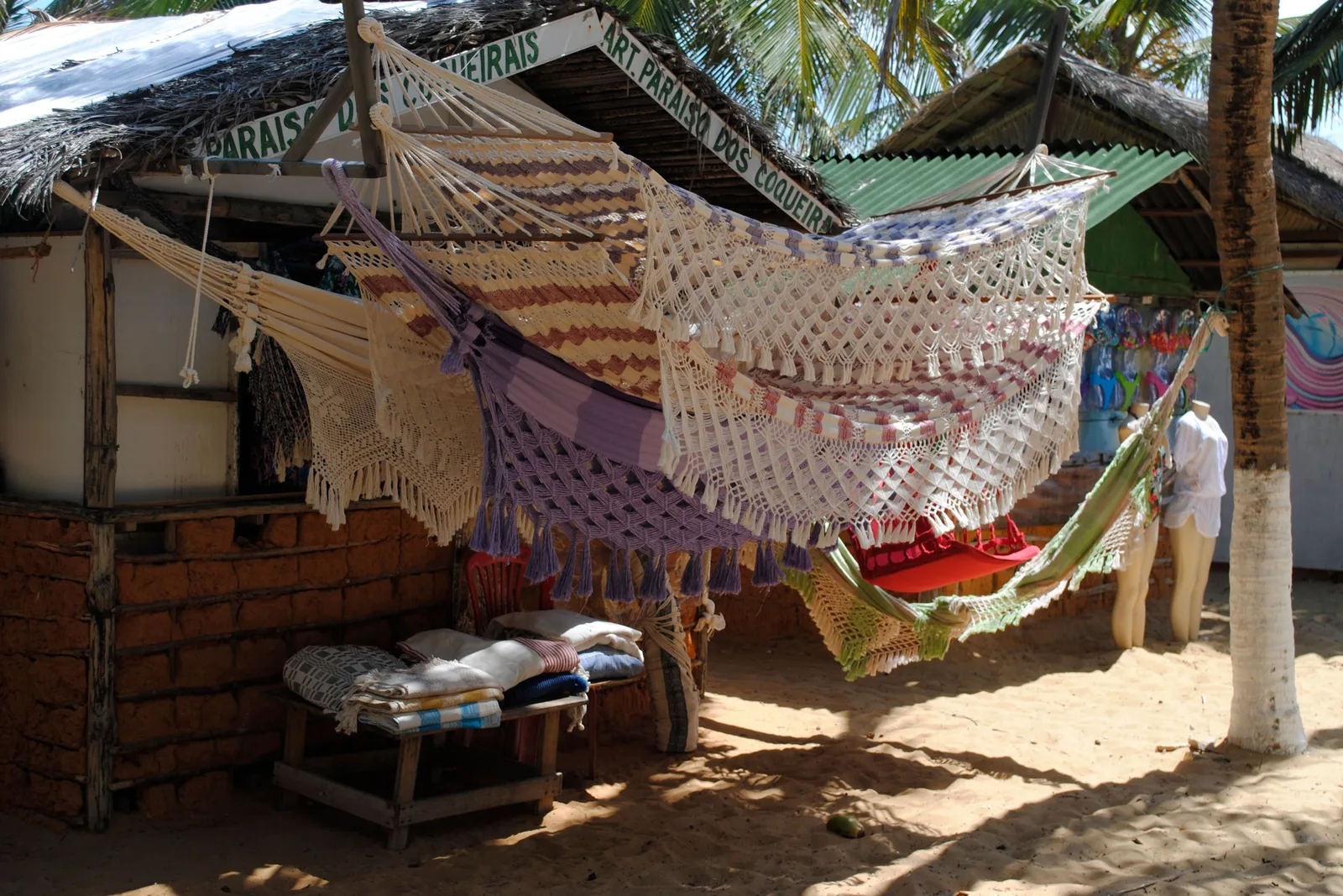 Hand-knotted hammocks in cream, lilac stripe and red hung for sale across the front of a thatched beach stall