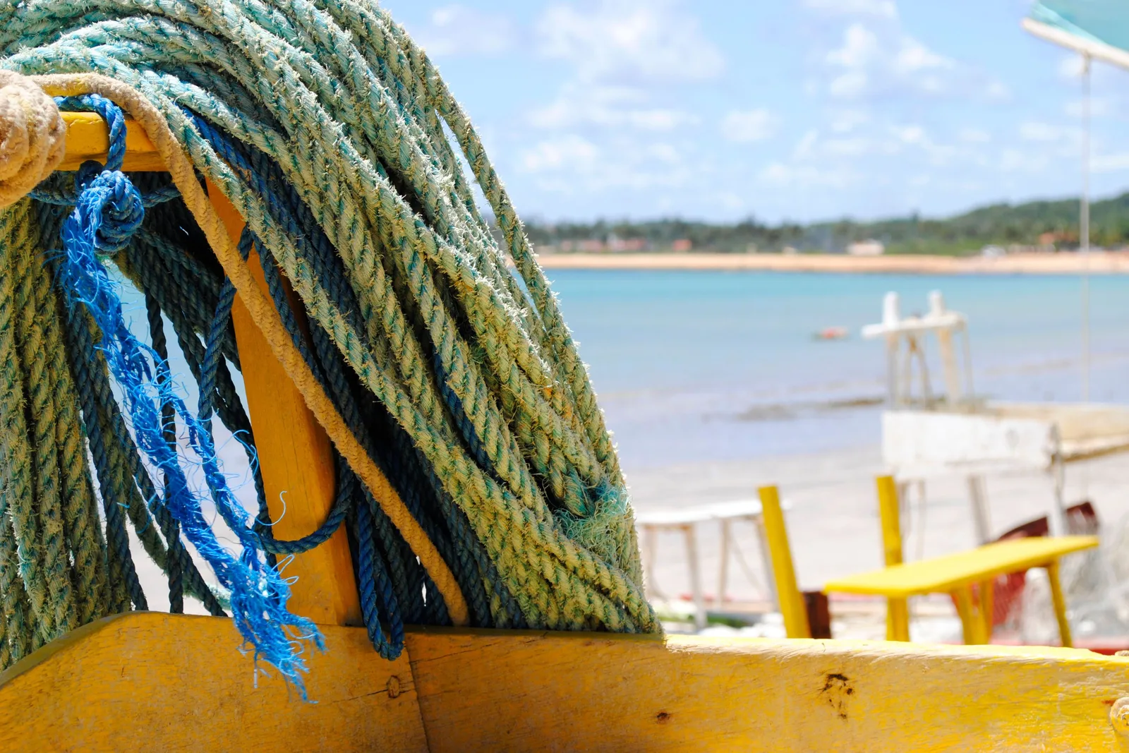 Coiled blue and green rope piled on the yellow deck of a jangada, with the beach and a row of chairs out of focus behind