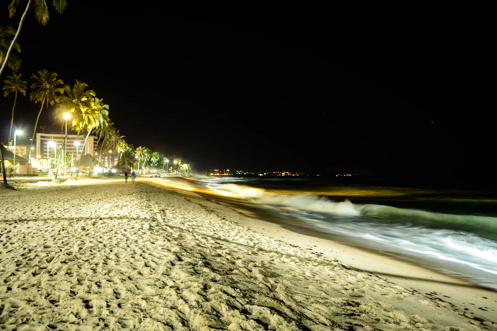 Jatiúca beach after dark in a long exposure: surf blurred to white across the sand and the lights of the promenade strung along behind