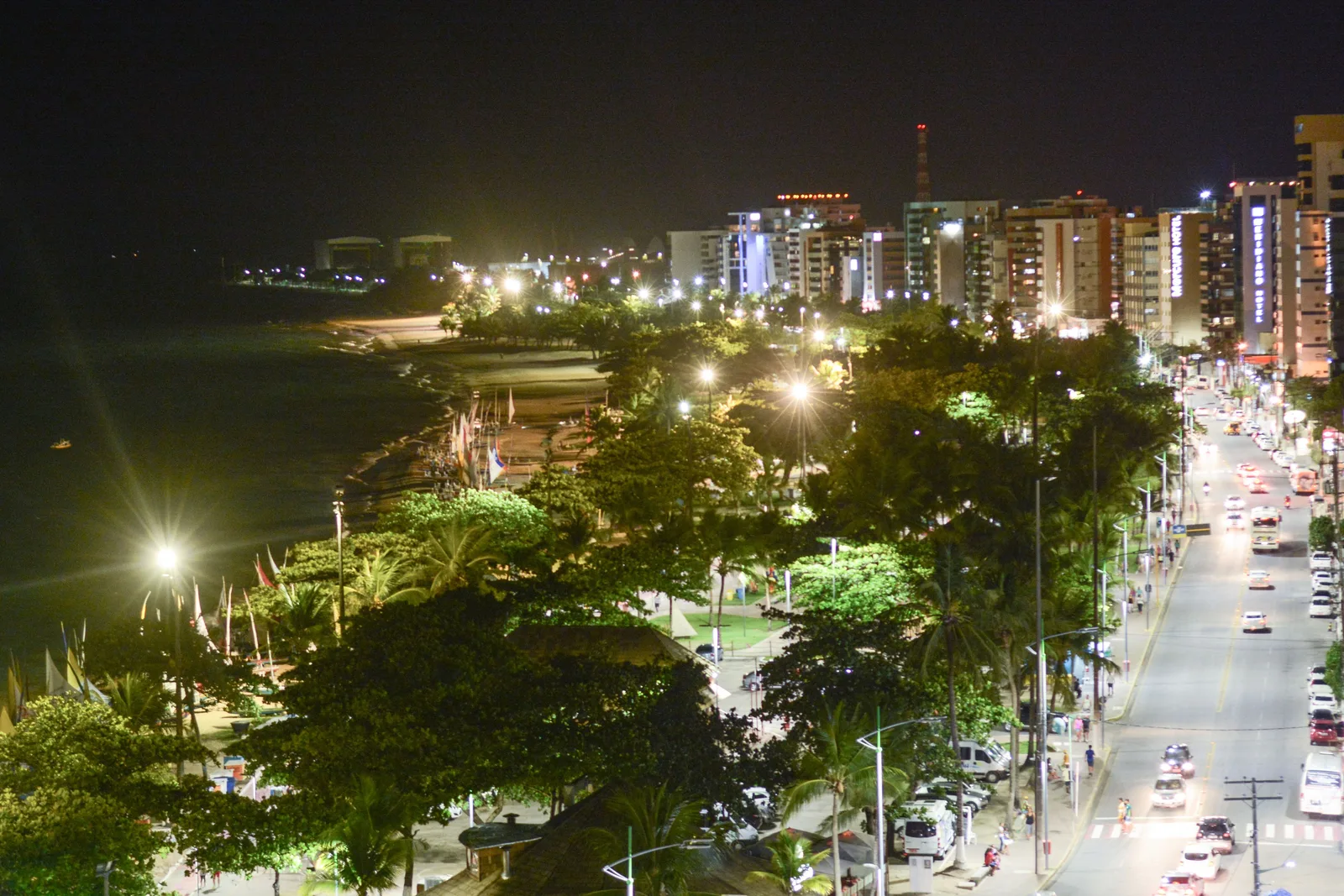 The Maceió seafront at night from above: traffic on a lit avenue curving past apartment blocks, with a band of floodlit broadleaf trees between the road and the dark sea