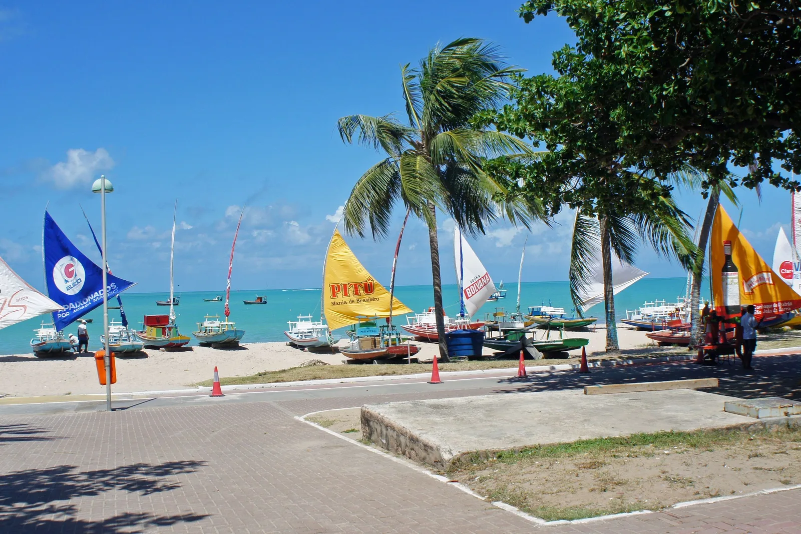 The Pajuçara seafront: jangadas drawn up on the sand beyond a paved plaza with their sails hoisted in blue, yellow, white and orange, palms leaning over them and turquoise sea behind