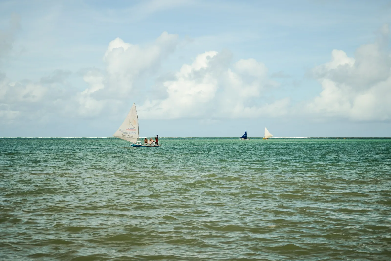 Three jangadas under sail on flat green water off Pajuçara, one close in with figures standing aboard and two more far out along the reef line, under a wide cloudy sky