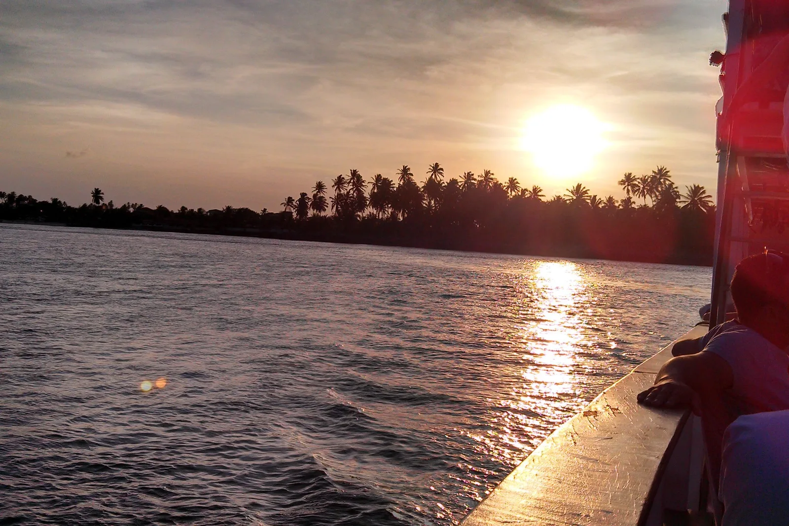 Sunset from a boat on the Mundaú lagoon: the sun low over flat water with a dark line of palms along the far shore
