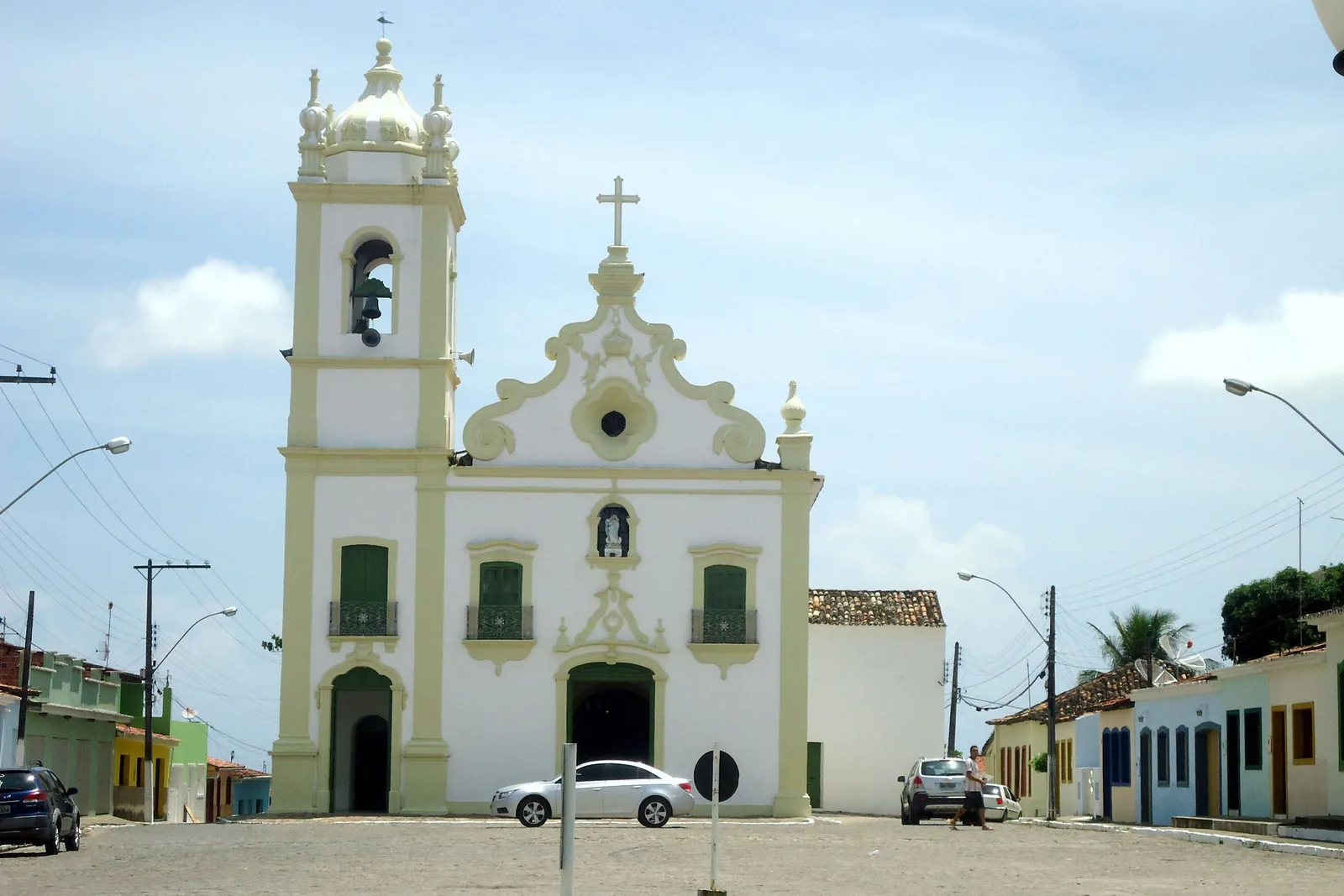 The parish church at Marechal Deodoro across its paved square: a cream and white baroque front with a single bell tower on the left, low colored colonial houses running away on either side