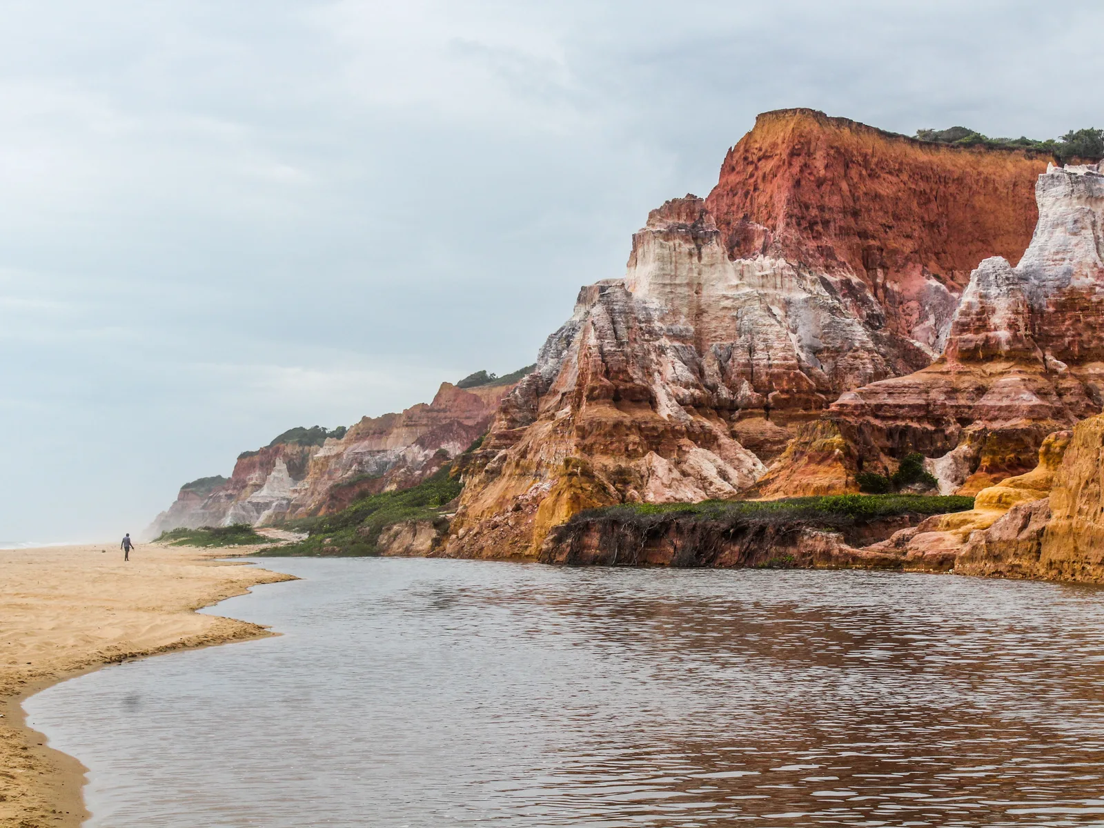 The banded red and cream cliffs at Praia do Gunga rising over a pale sand spit, with the lagoon channel running out to the sea in the foreground and a single figure walking the sand