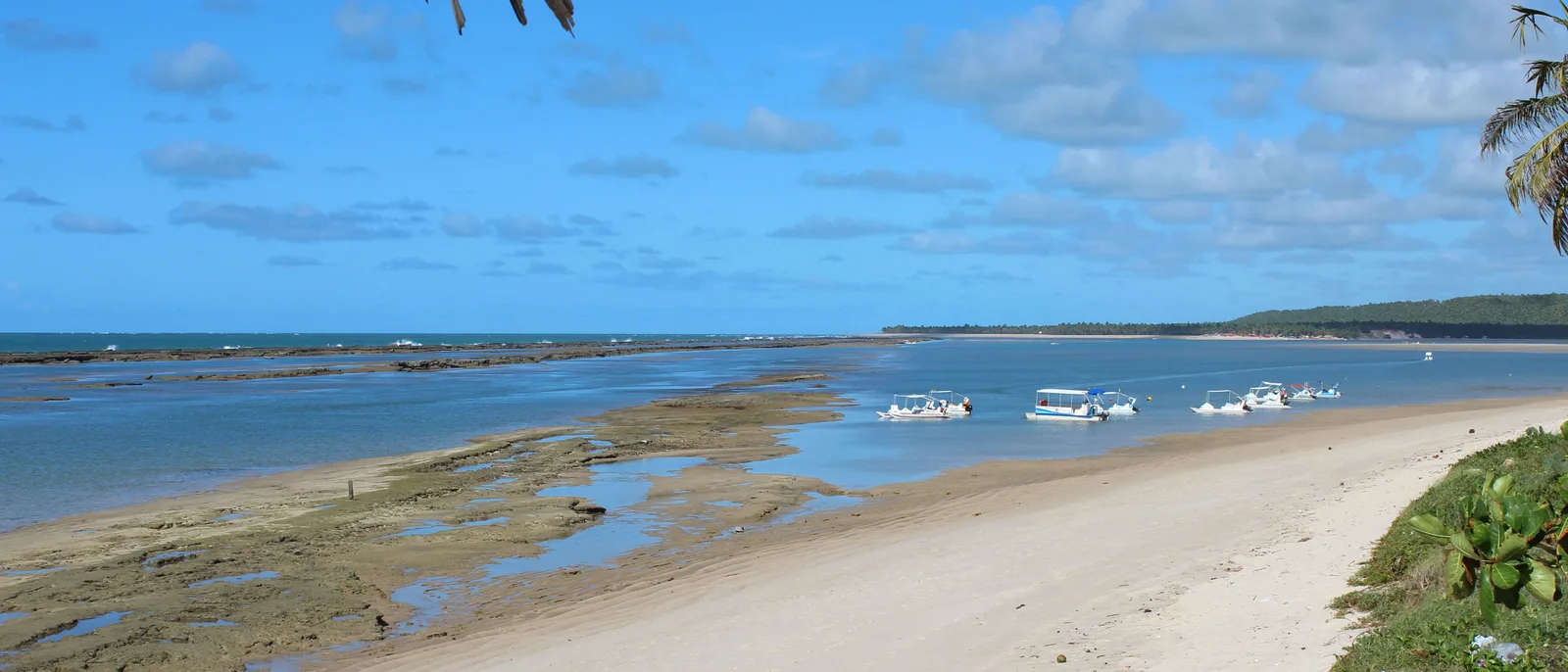 The beach at Barra de São Miguel in a wide frame: boats moored on flat water inside a reef line, pale sand and coconut palms along the shore