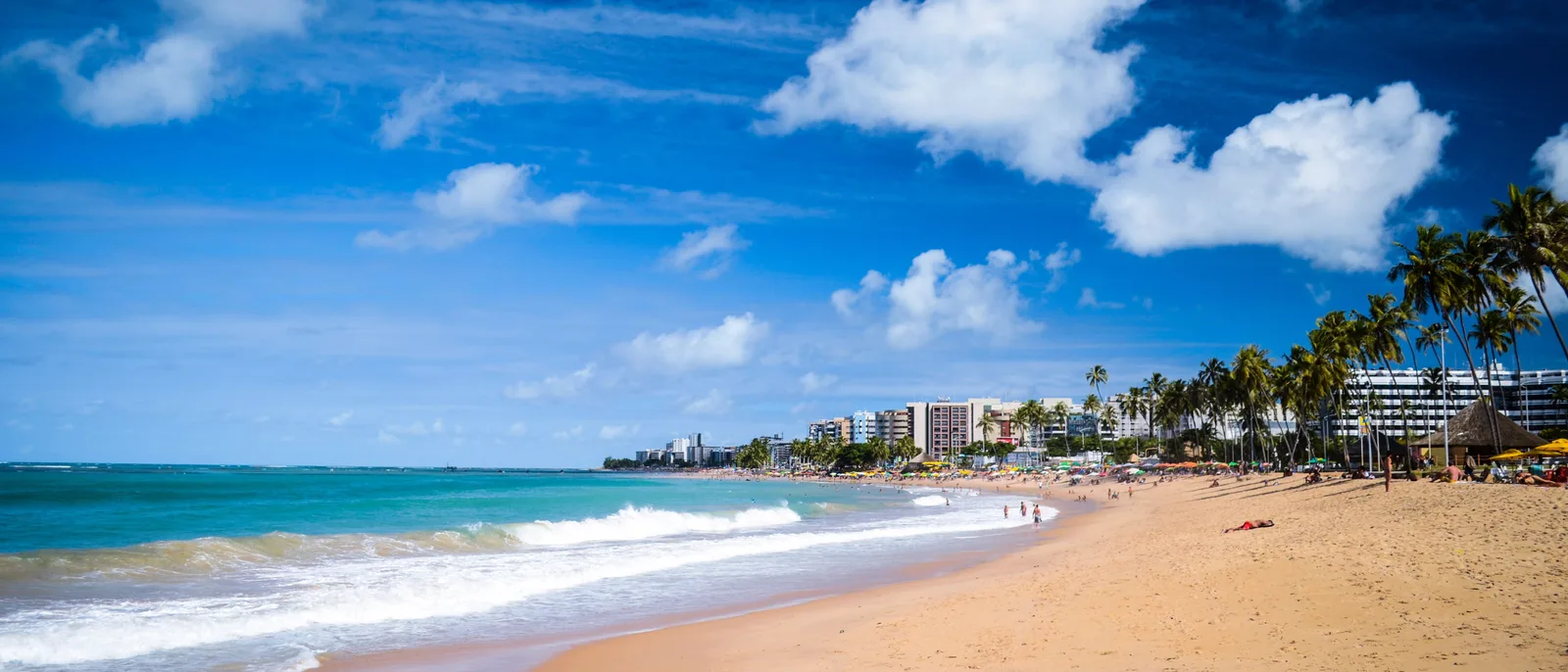 Jatiúca beach in a wide frame: surf breaking on an open stretch of sand with palms and the buildings of the seafront running away into the distance