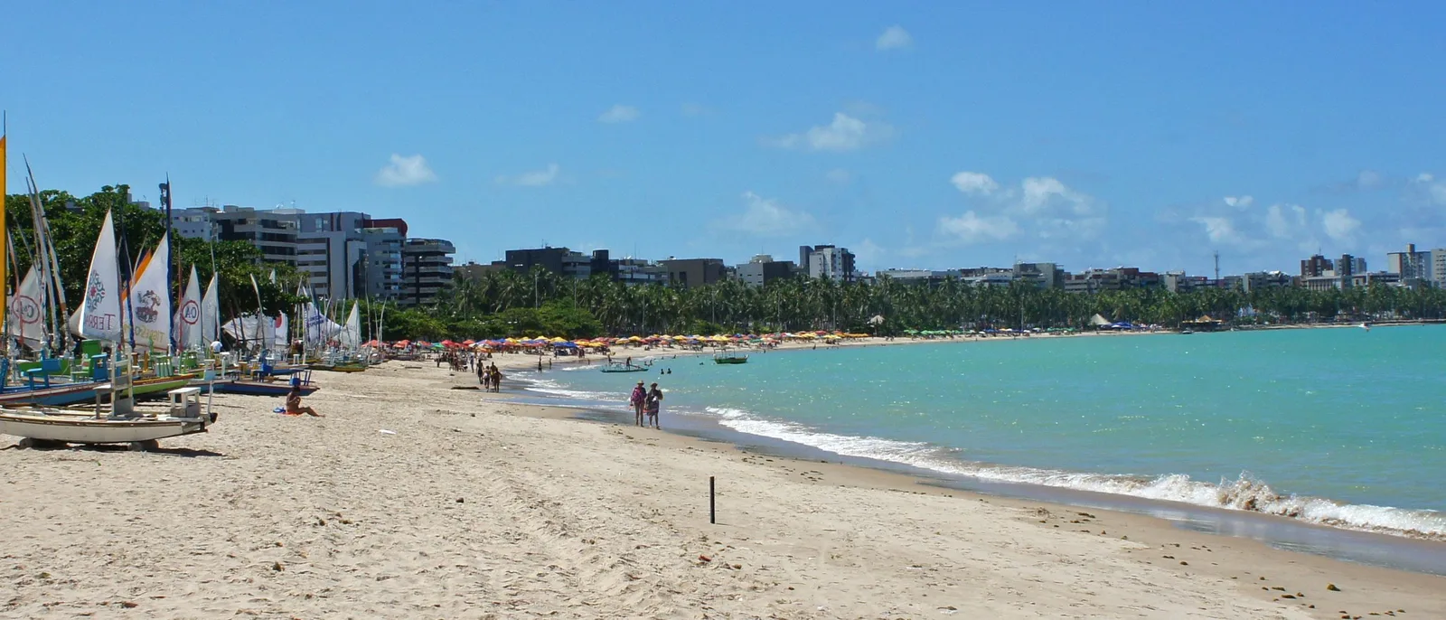 The Pajuçara seafront in a wide panorama: green shallow water, a broad strip of sand and a continuous line of apartment towers behind the palms