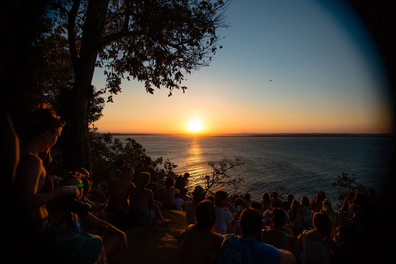 A crowd sitting along a grassy clifftop at Morro de São Paulo watching the sun go down over the water, boats small on the horizon below them